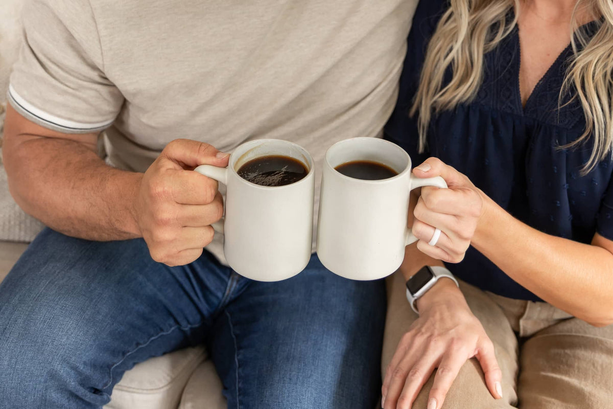 Close-up of Elo Coffee Co. founders Andrew and Emily Meade's hands holding two white mugs filled with black coffee, sitting side by side on a couch.