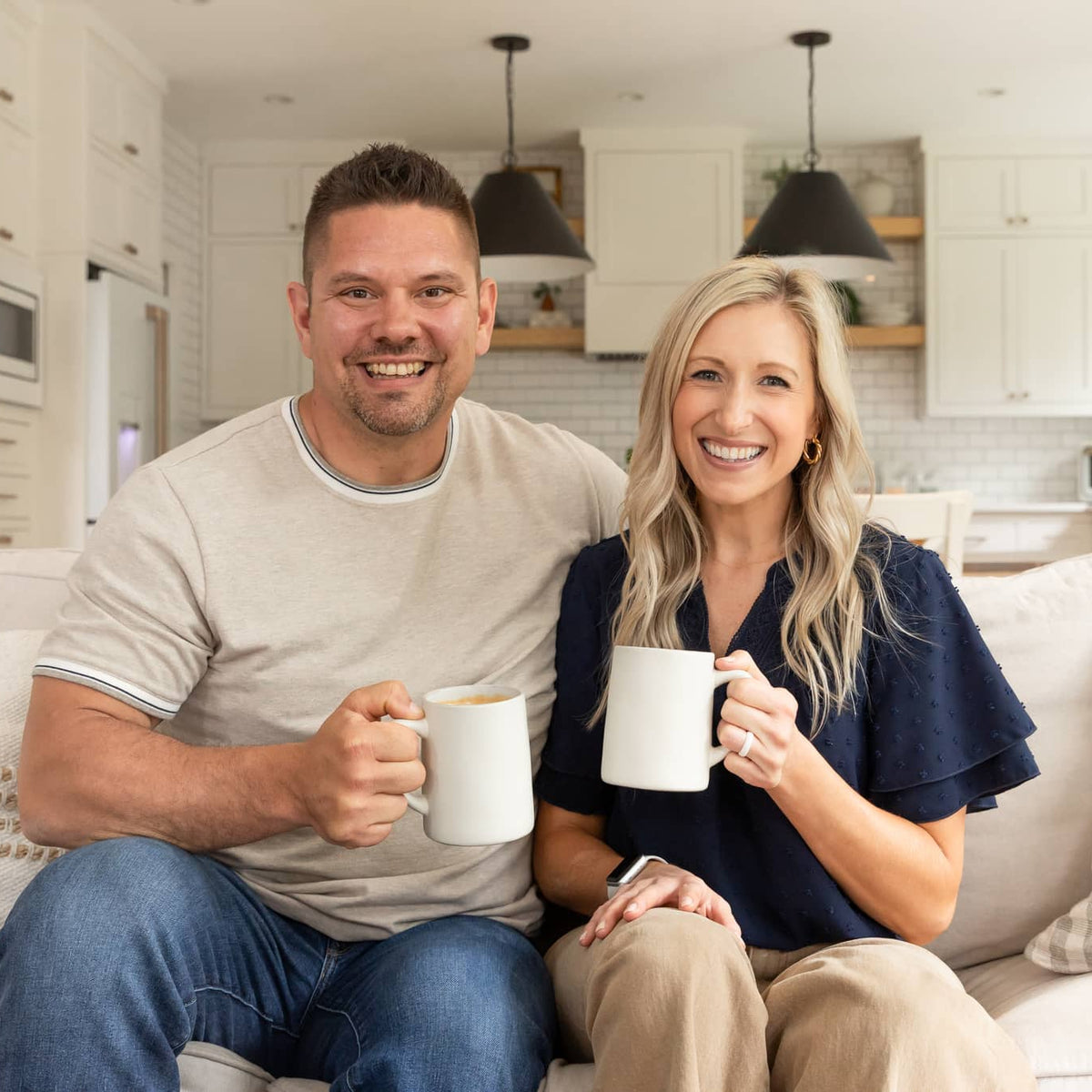 Elo Coffee Co. founders Andrew and Emily Meade sitting together on a couch at home, each holding a white coffee mug and smiling at the camera, with a bright modern kitchen visible in the background.